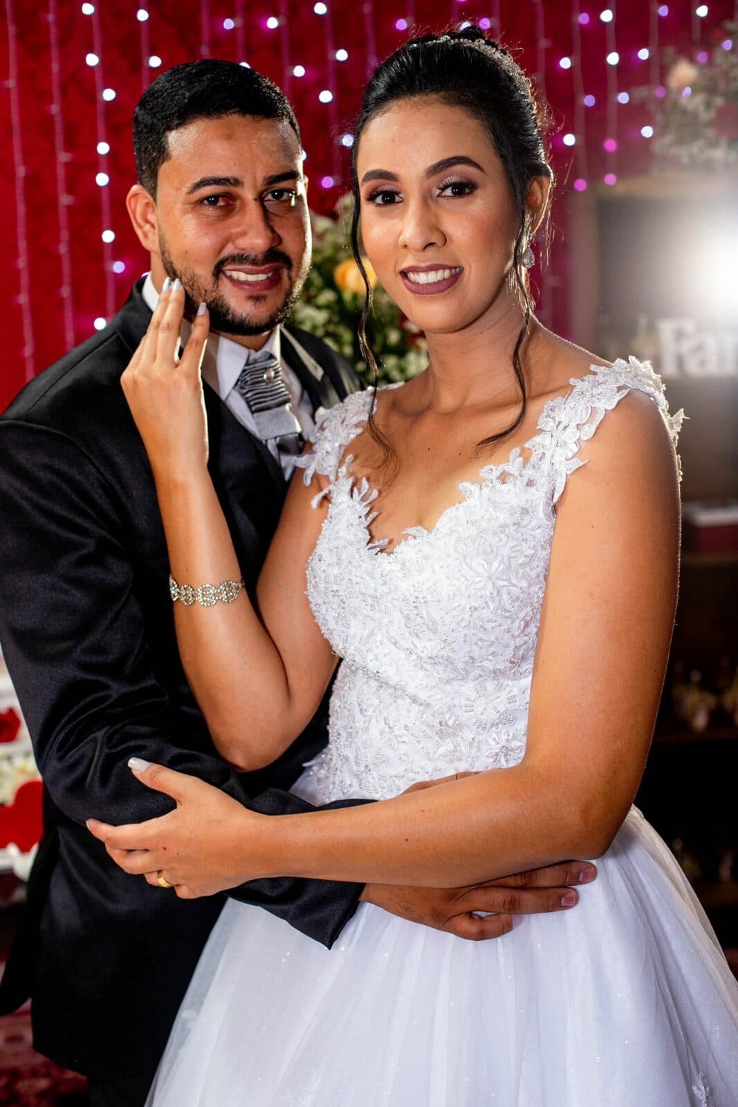 Retrato do casal abraçado diante de uma decoração vermelha com luzes.